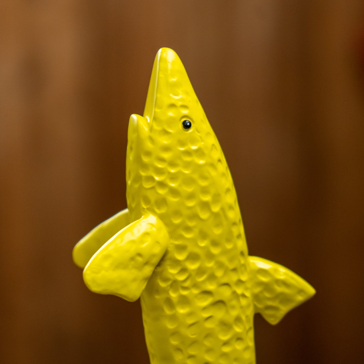 A close-up of the Merchtank Yellow Shark Tap Handle, a neon yellow, textured figurine with an open mouth and outstretched fins, set against a brown blurred background.