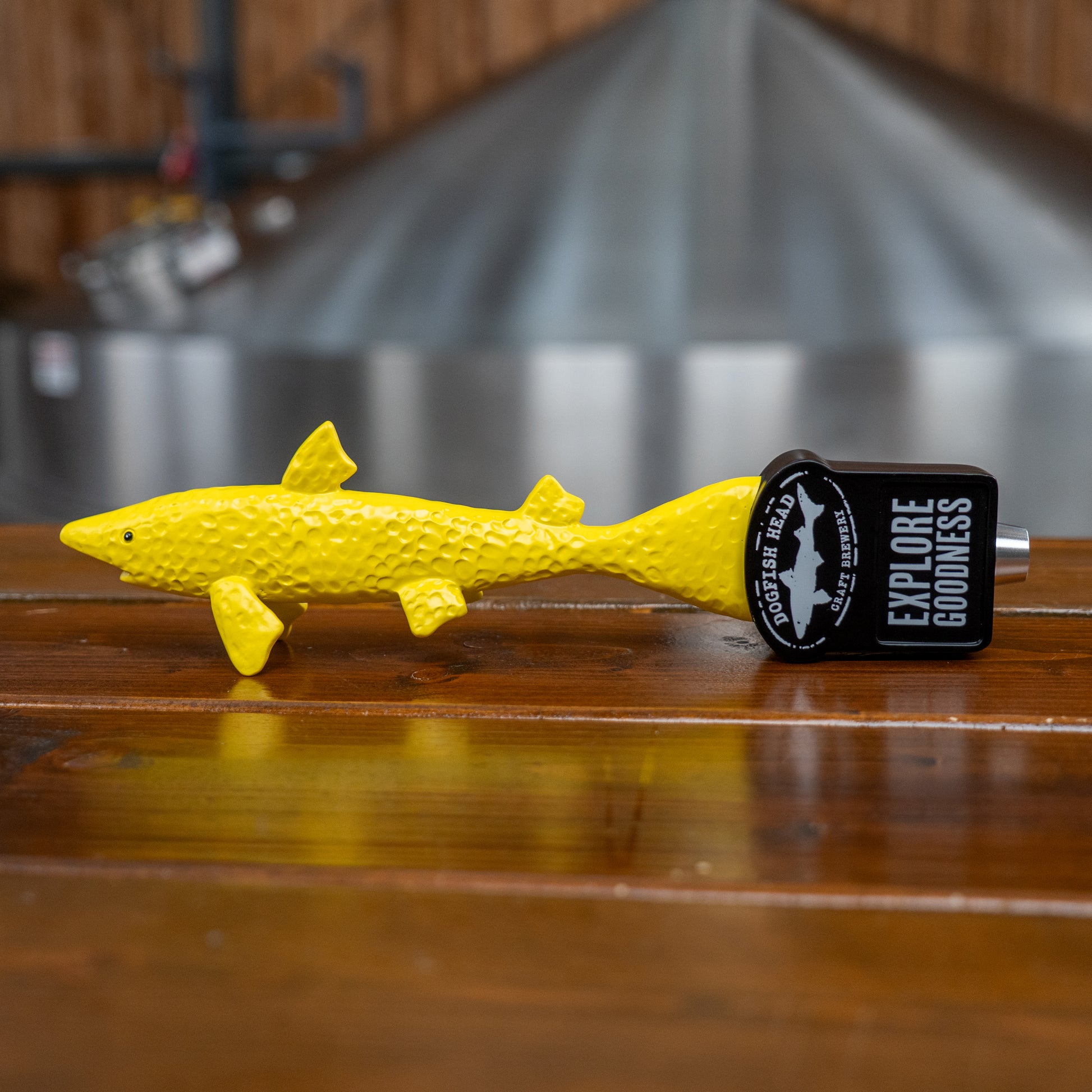 A neon yellow fish-shaped Yellow Shark Tap Handle by Merchtank is attached to a beer tap reading EXPLORE GOODNESS, sitting on a wooden bar alongside other tap handles with a blurred brewery in the background.