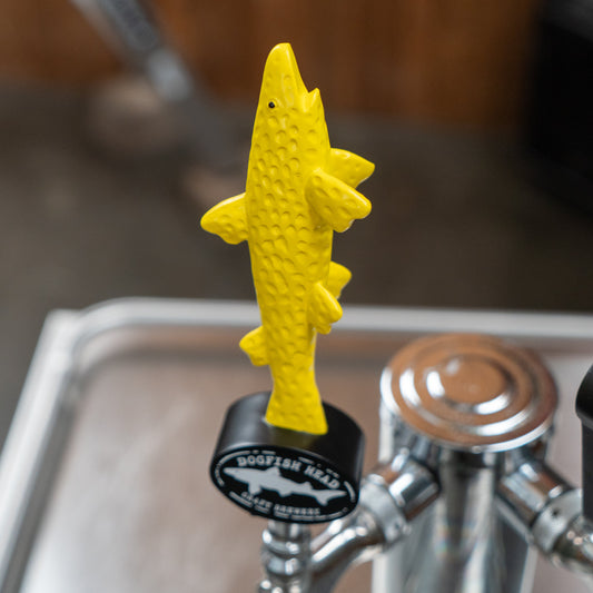 A Yellow Shorty Tap Handle from Merchtank, shaped like a fish, is attached to a draft beer dispenser pouring fresh beer, with blurred bar equipment in the background.