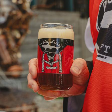 Glass shown being held by someone and filled with beer. Front view of can glass features Dogfish Head shark and shield logo and is surrounded by a red hockey jersey.