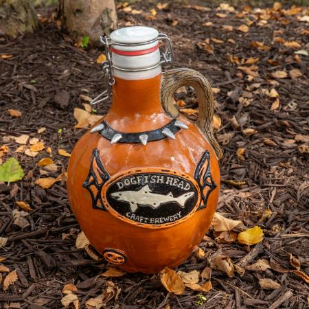 Close-up of the orange growler. Featured is a black collar on the neck of the growler with studs. The Dogfish Head shark and shield logo is centered on the front with bat wings on it. A small orange pumpkin is featured on the bottom right.