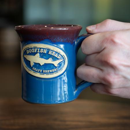 Shown being held, is a blue mug with a red drip glaze on top. The front features a white Dogfish Head shark and shield logo.