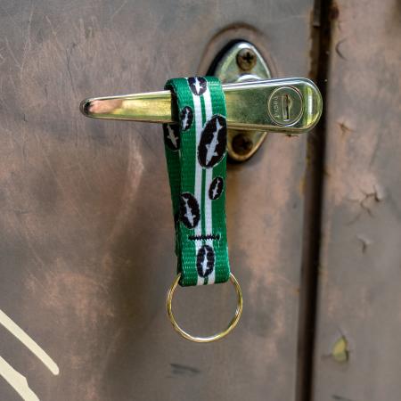 Green keychain has 2 white lines going down the middle and is covered in small black and white Dogfish Head shark and shield logos. Image shows keychain hanging on a door handle.