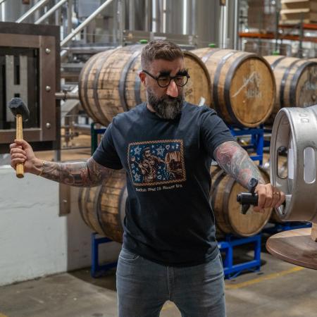 Shown by a person wearing, front view of the shirt features Jon Langford's retro label artwork on a blue background, along with writing "Dogfish Head 75 Minute IPA". 