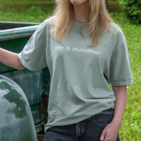  A person wears a light sage green t-shirt with the text "beer & benevolence" printed across the front in white. Only the torso is visible, standing next to a vintage green truck.