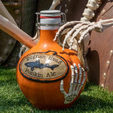 Orange growler with Dogfish head logo centered with writing "Dogfish Head Punkin Ale". Growler handle is brown made to look like a tree branch. Skeleton hand is featured on the bottom of the growler.