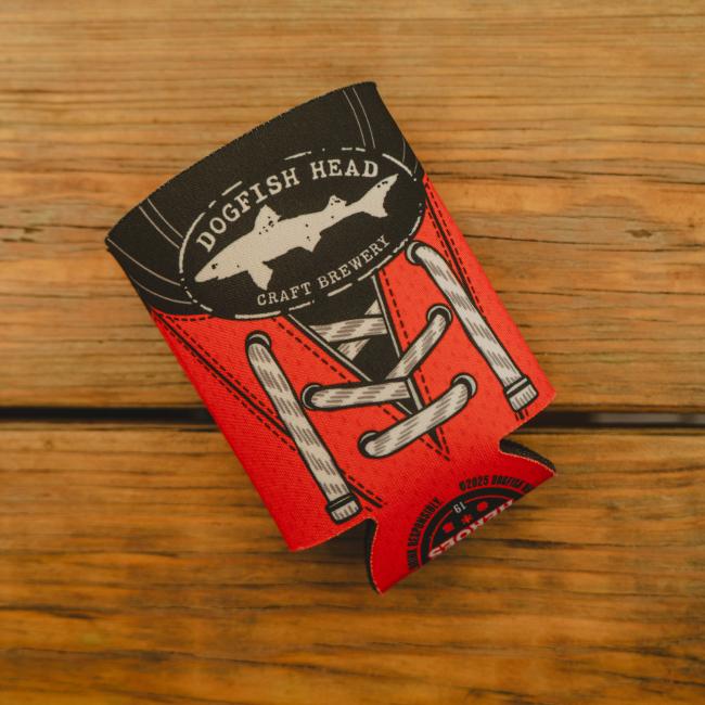 View of can cooler laid flat on a table. Cooler features Dogfish Head shark and shield logo and is made to look like a red hockey jersey.