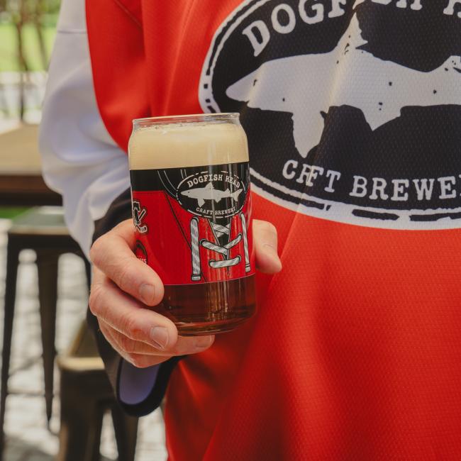 Glass shown being held by someone and filled with beer. Front view of can glass features Dogfish Head shark and shield logo and is surrounded by a red hockey jersey.