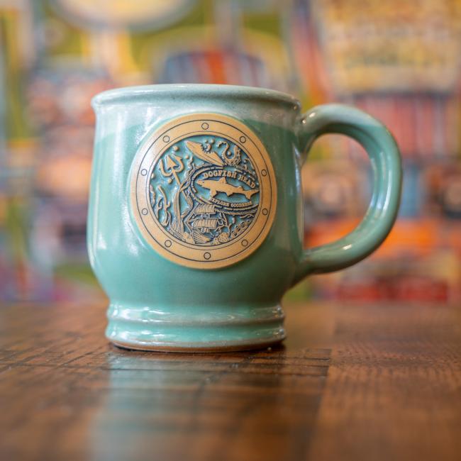 Mint colored mug which features imagery of a port hole with sea articles and the Dogfish Head shark and shield logo inside it.