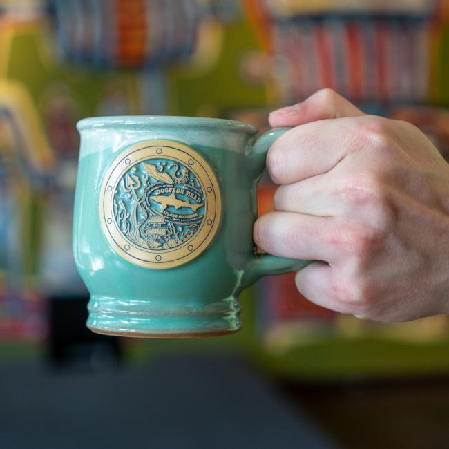 Image of mug being held up. Mint colored mug features imagery of a port hole with sea articles and the Dogfish Head shark and shield logo inside it.