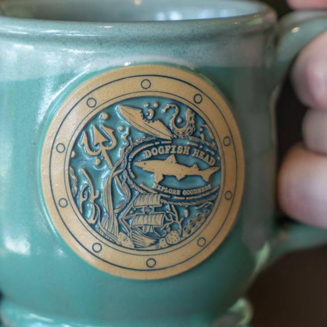 Close-up of the port hole on the front of the mug. Image of mug being held up. Features sea articles and the Dogfish Head shark and shield logo inside it.