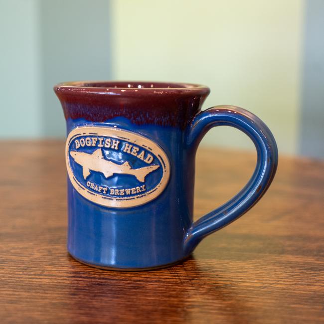 A blue mug with a red drip glaze on top. The front features a white Dogfish Head shark and shield logo.