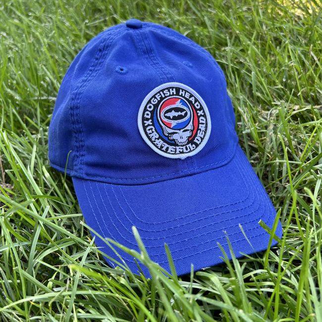 Front view of royal blue hat shown laid on grass. Hat features Dogfish Head x Grateful Dead collaboration logo emblem centered to front of the hat.