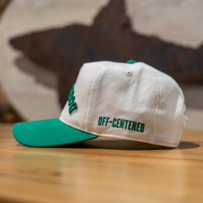 A cream colored five panel hat with a green brim and green embroidery that says