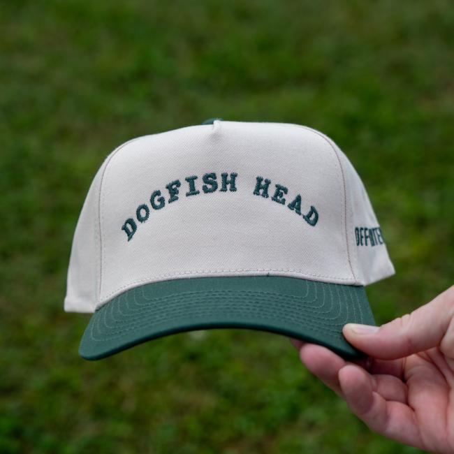 Shown held, is the front view of a white hat with a green brim. Featured arched on the front is 'Dogfish Head' written in green.