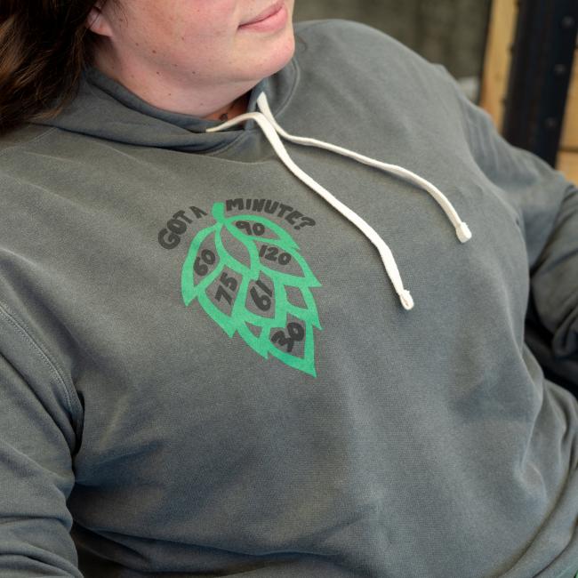 Close-up of front view of grey hoodie being modeled. Features a green hop cone graphic with numbers '60', '90', '75', '120', '61' and '30' inside it, and the text 'GOT A MINUTE?' above it, on the right side of chest.