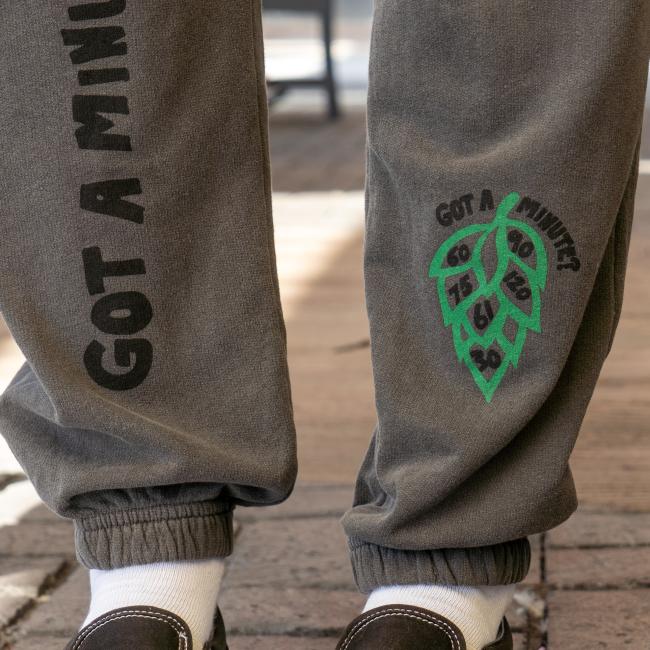 Close-up of the bottom of the sweatpants, the left features a green hop cone graphic with numbers '60', '90', '75', '120', '61' and '30' inside it, and the text 'Got A Minute?' above it, and right has writing 