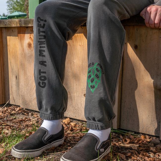 Close-up of the bottom of the sweatpants, the left features a green hop cone graphic with numbers '60', '90', '75', '120', '61' and '30' inside it, and the text 'Got A Minute?' above it, and right has writing 