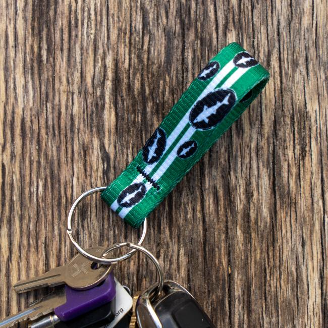Green keychain has 2 white lines going down the middle and is covered in small black and white Dogfish Head shark and shield logos. Image shows keychain in a person's hand with keys hanging on it.