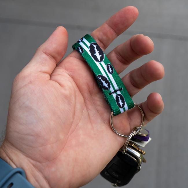 Green keychain has 2 white lines going down the middle and is covered in small black and white Dogfish Head shark and shield logos. Image shows keychain in a person's hand with a car key hanging on it.