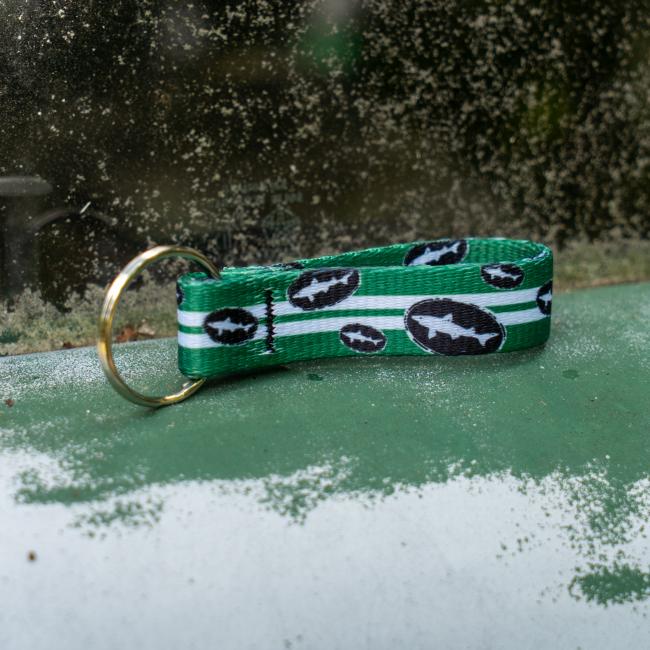 Green keychain has 2 white lines going down the middle and is covered in small black and white Dogfish Head shark and shield logos. Image shows keychain laid on its side.