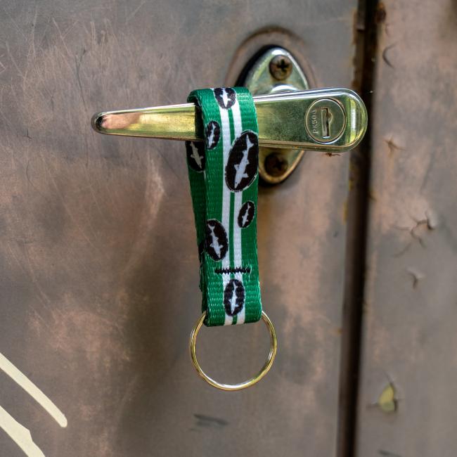 Green keychain has 2 white lines going down the middle and is covered in small black and white Dogfish Head shark and shield logos. Image shows keychain hanging on a door handle.