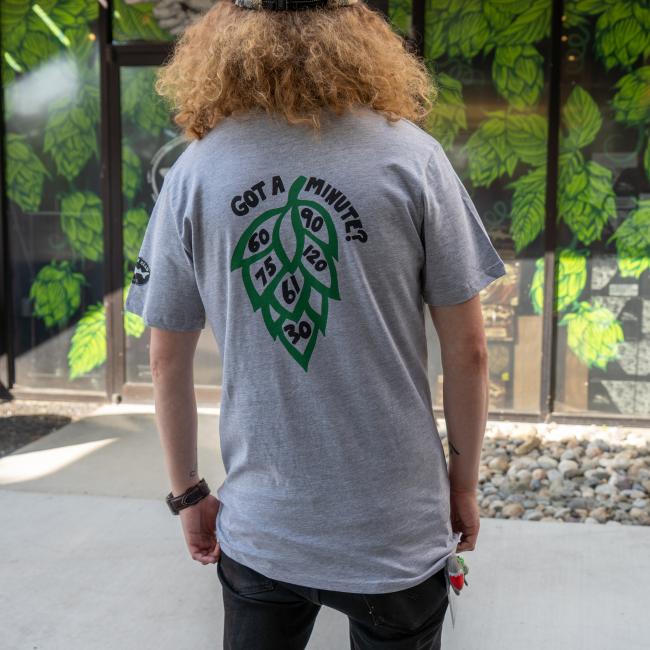 Tee being modeled, back features a large green hop cone graphic with numbers '60', '90', '75', '120', '61' and '30' inside it, and the text 'GOT A MINUTE?' above it.