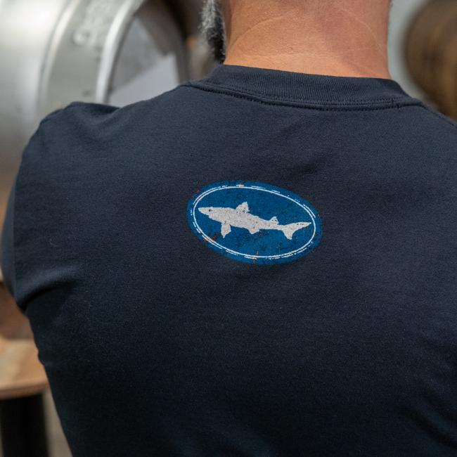 Close-up of back of shirt on a person, featuring a blue Dogfish Head shark and shield logo by the neck area.