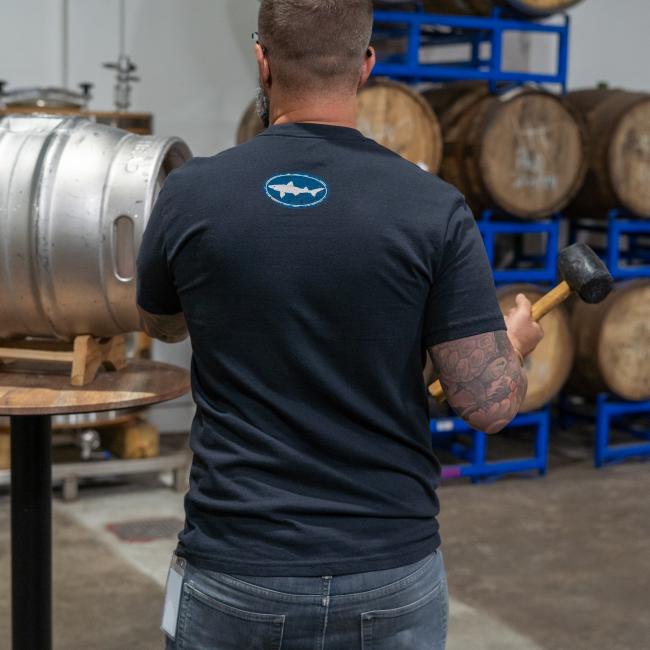 Back of shirt on a person, featuring a blue Dogfish Head shark and shield logo by the neck area.